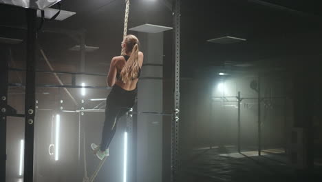 Full-length-wide-angle-shot-of-a-woman-performing-rope-climbs-at-the-gym.-Athletics-healthy-composition.-Cross-fitness.-Determined-athletic-woman-moving-up-the-rope-in-a-gym.